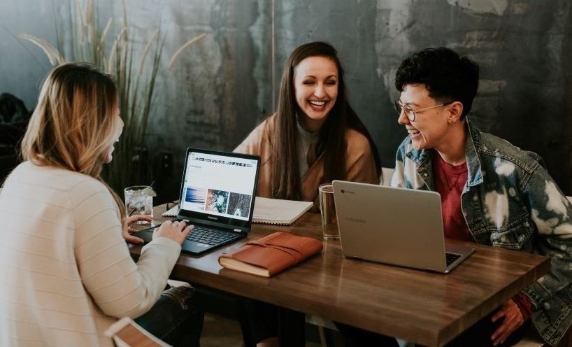 Three people are sitting at a table smiling and laughing. There are two laptops on the table.