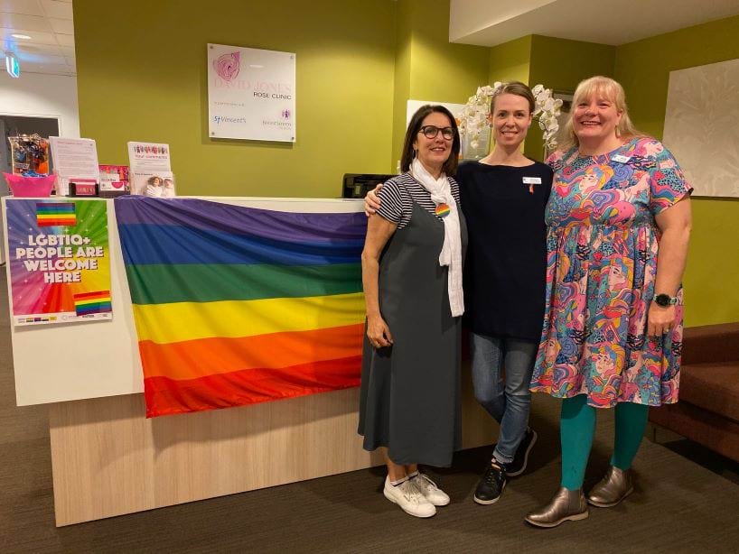 Three people similing and standing in front of a rainbow flag with a sign saying LGBTIQ+ people are welcome here. 