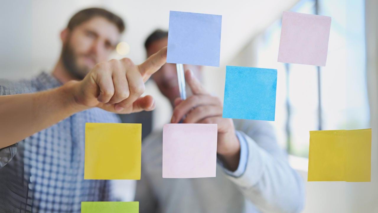 You are looking through a see-through board, which has multiple post it notes stuck on it. On the other side of the board, there are 2 people pointing towards you at a post it note in the middle of the board.