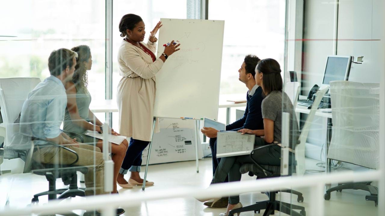 You are looking through a glass window with white panes at 5 people who seem to be in a work meeting. In the middle is a person standing up and talking, while signalling their hands towards a whiteboard. They are holding a marker in their hand. Sitting on either side of them are 2 people looking towards the whiteboard. Some of them are holding books or an iPad in their hands.