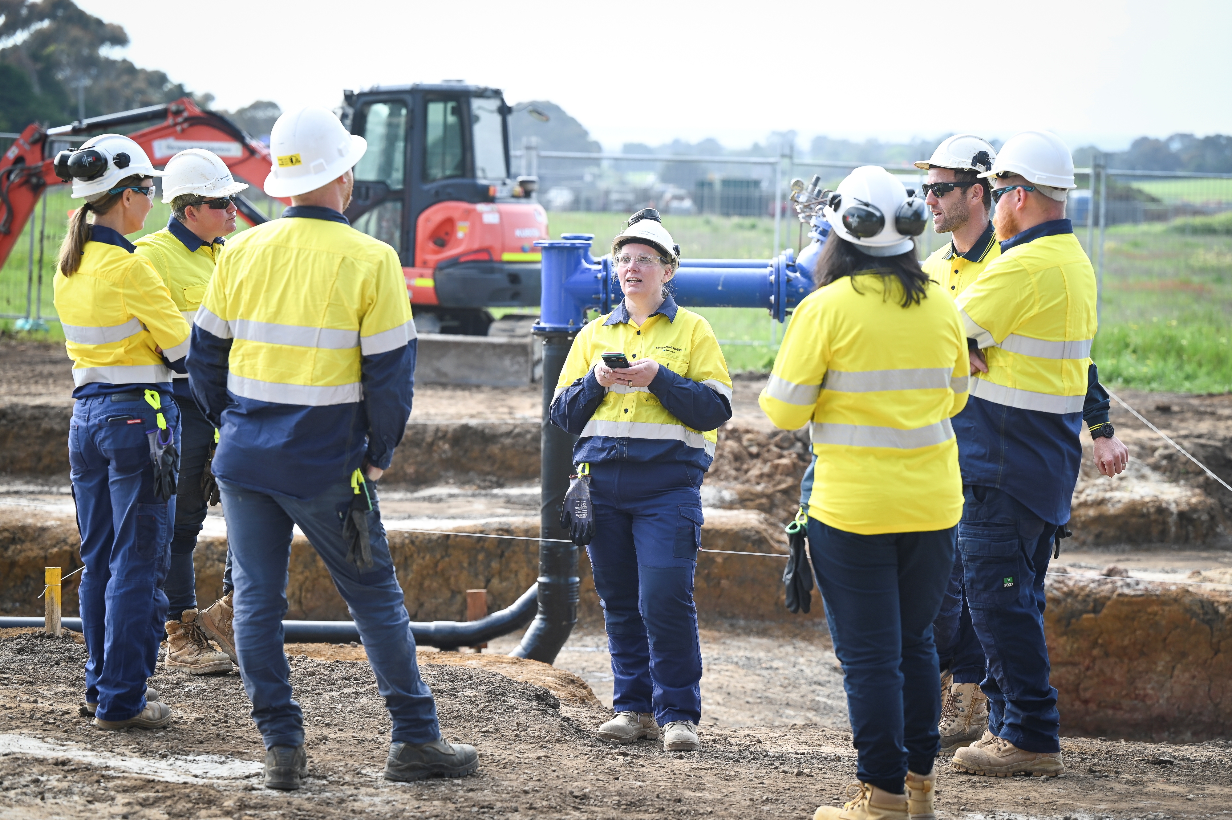 A group of construction workers, wearing yellow high-visibility safety gear and hard hats, stand in a circle having a discussion at an outdoor worksite with excavation equipment and pipes in the background.