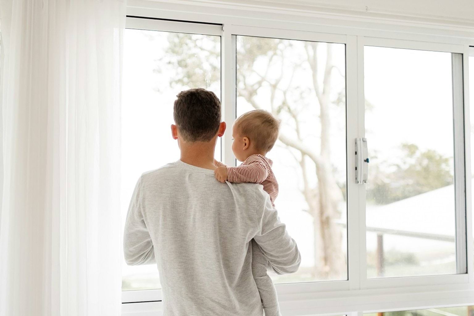 A father holding his young child while looking out a large window during the day. The child is resting their hand on the father's shoulder.