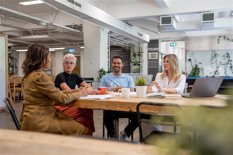 Four professionals having a meeting around a wooden table in a modern office space with exposed ceiling, white pillars, and plants. The group includes people of varying ages engaged in discussion, with laptops, notebooks, and coffee cups on the table.